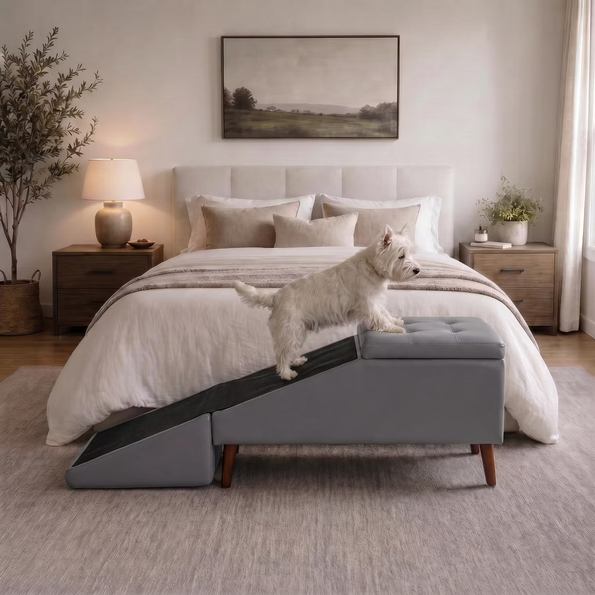 A small white dog stands on a gray pet ramp positioned in front of a bed in a softly styled bedroom. The ramp has a dark gray textured surface and a matching gray tufted rectangular end, with wooden legs.