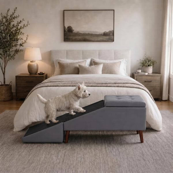 A small white dog stands on a gray pet ramp positioned in front of a bed in a softly styled bedroom. The ramp has a dark gray textured surface and a matching gray tufted rectangular end, with wooden legs.